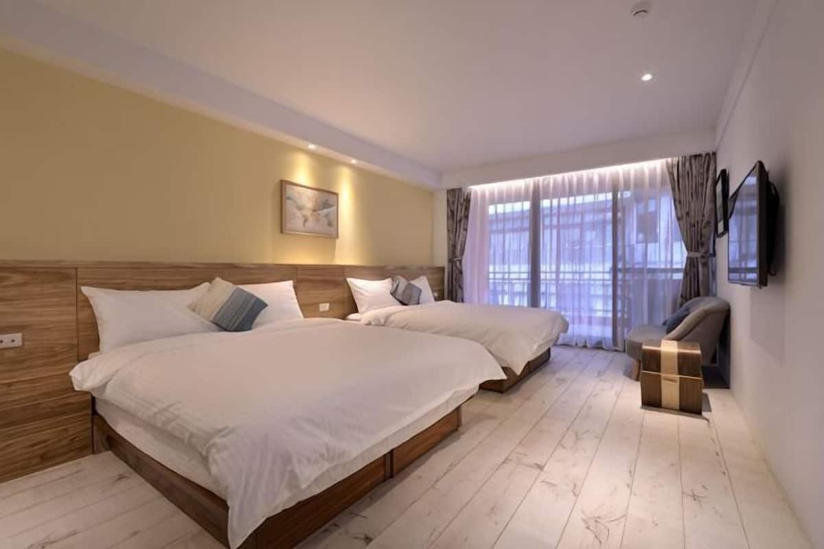 A hotel room with two beds, a flat-screen TV, a wooden headboard, and a large window with curtains.