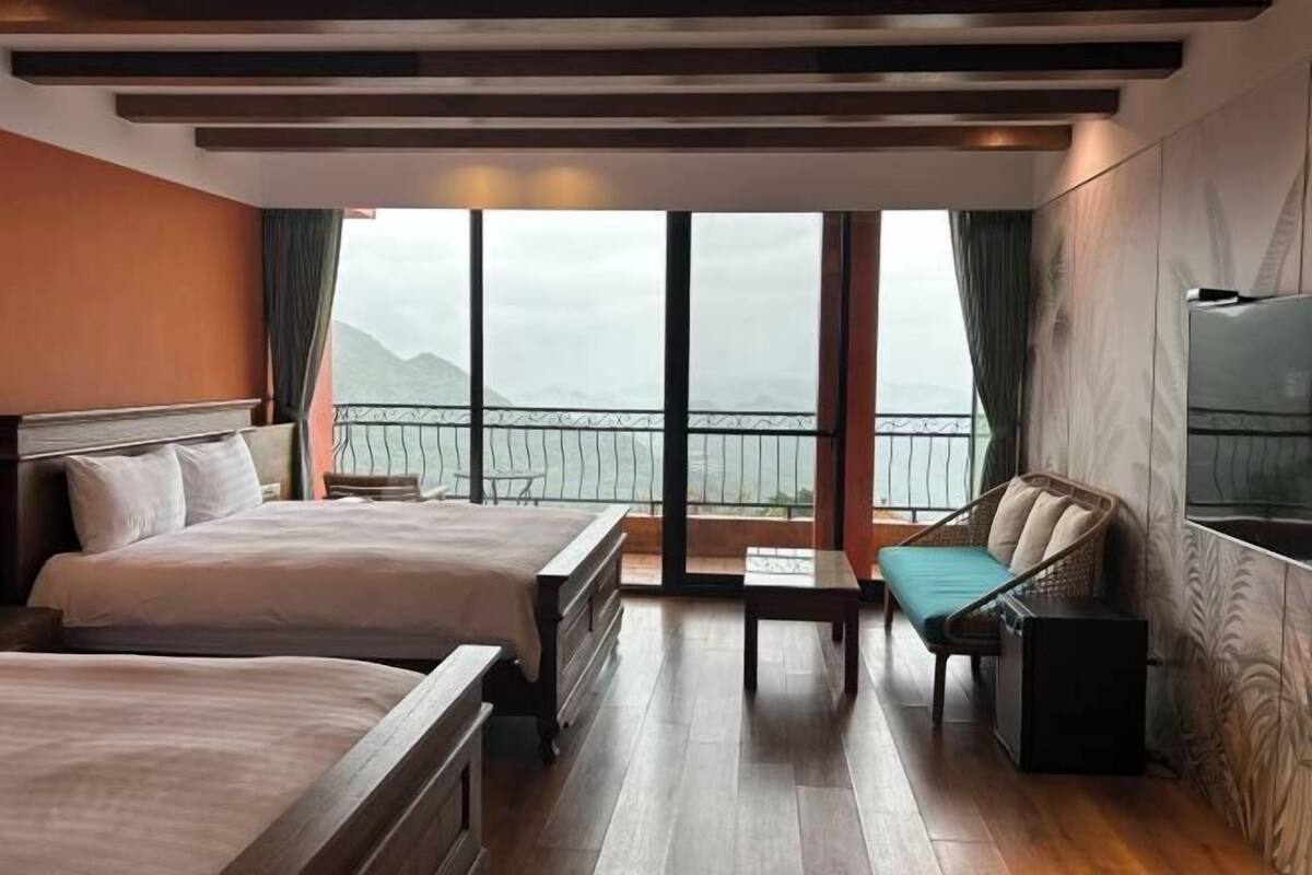 A hotel room with two beds, a small table, a TV, and a balcony with a view of the sea.