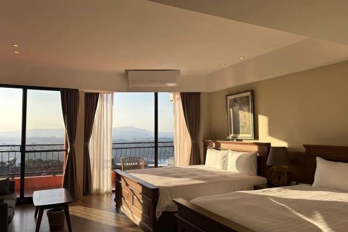 A hotel room with two beds, a desk, and a view of mountains through large windows.