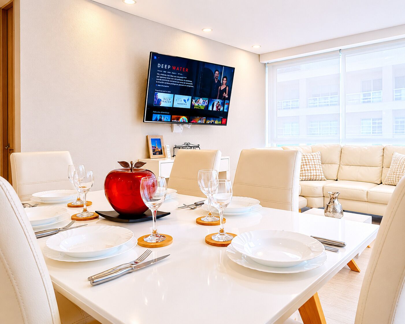 A dining area with a white table set for four, a red apple centerpiece, and a flat-screen TV mounted on the wall displaying a movie.