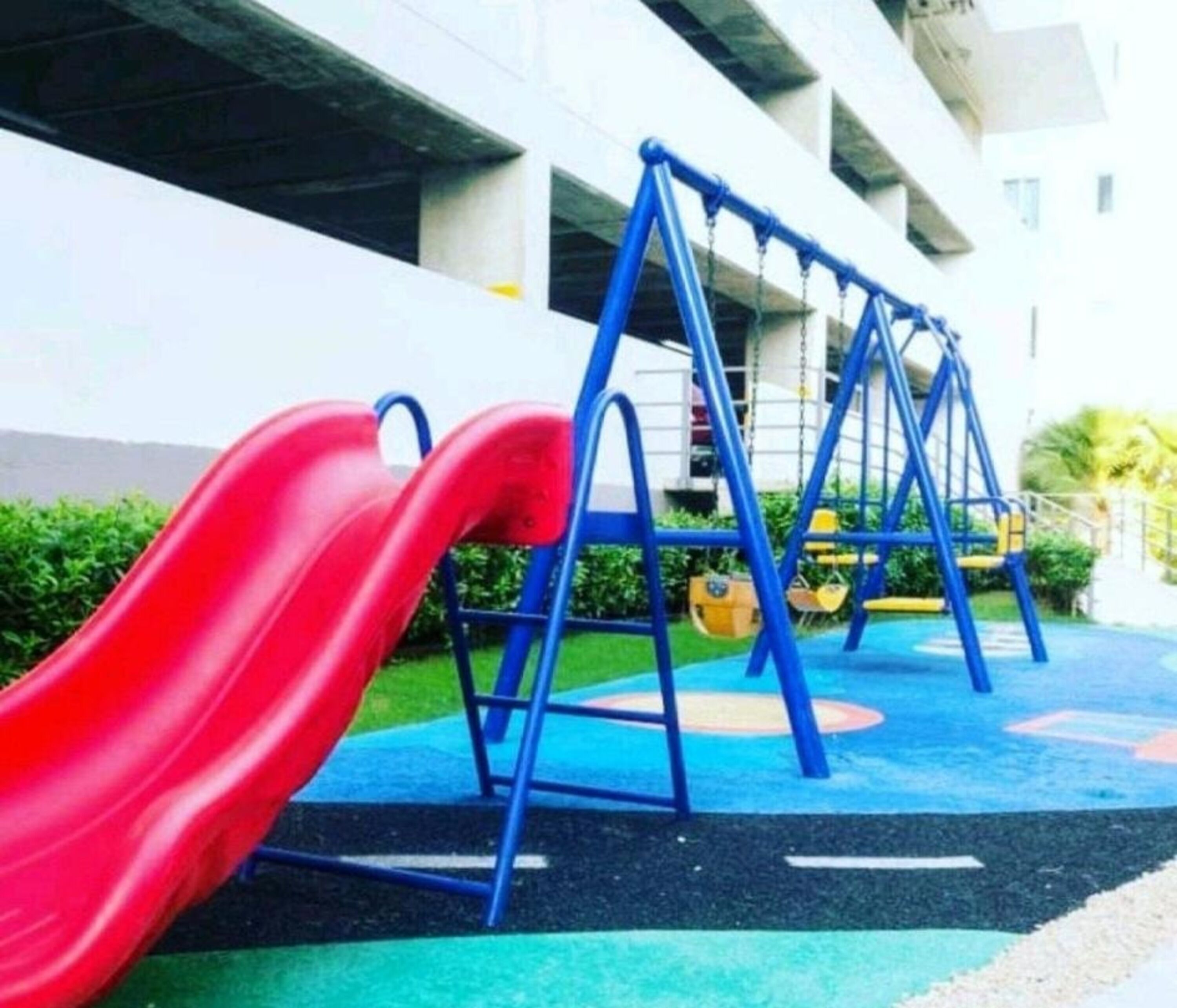 A playground with a red slide and blue swings in front of a building.