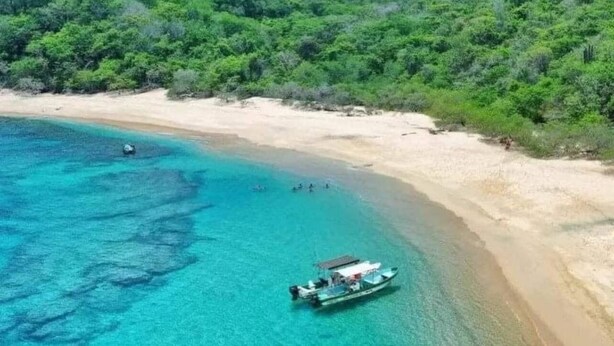 Una playa con aguas turquesas cristalinas, un bote y una colina boscosa al fondo.