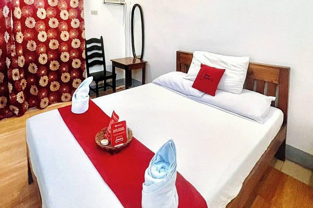 Ein ordentlich gemachtes Bett mit einer roten Fußmatte, ein kleiner Tisch mit einem Korb und einer Kerze sowie ein Stuhl.