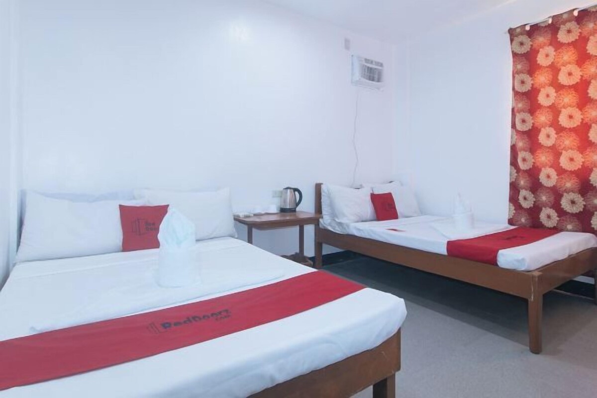 Habitación de hotel con dos camas individuales, cada una con ropa blanca de cama y detalles rojos. Hay una mesita con una tetera y una taza, y una cortina roja y blanca en la ventana.