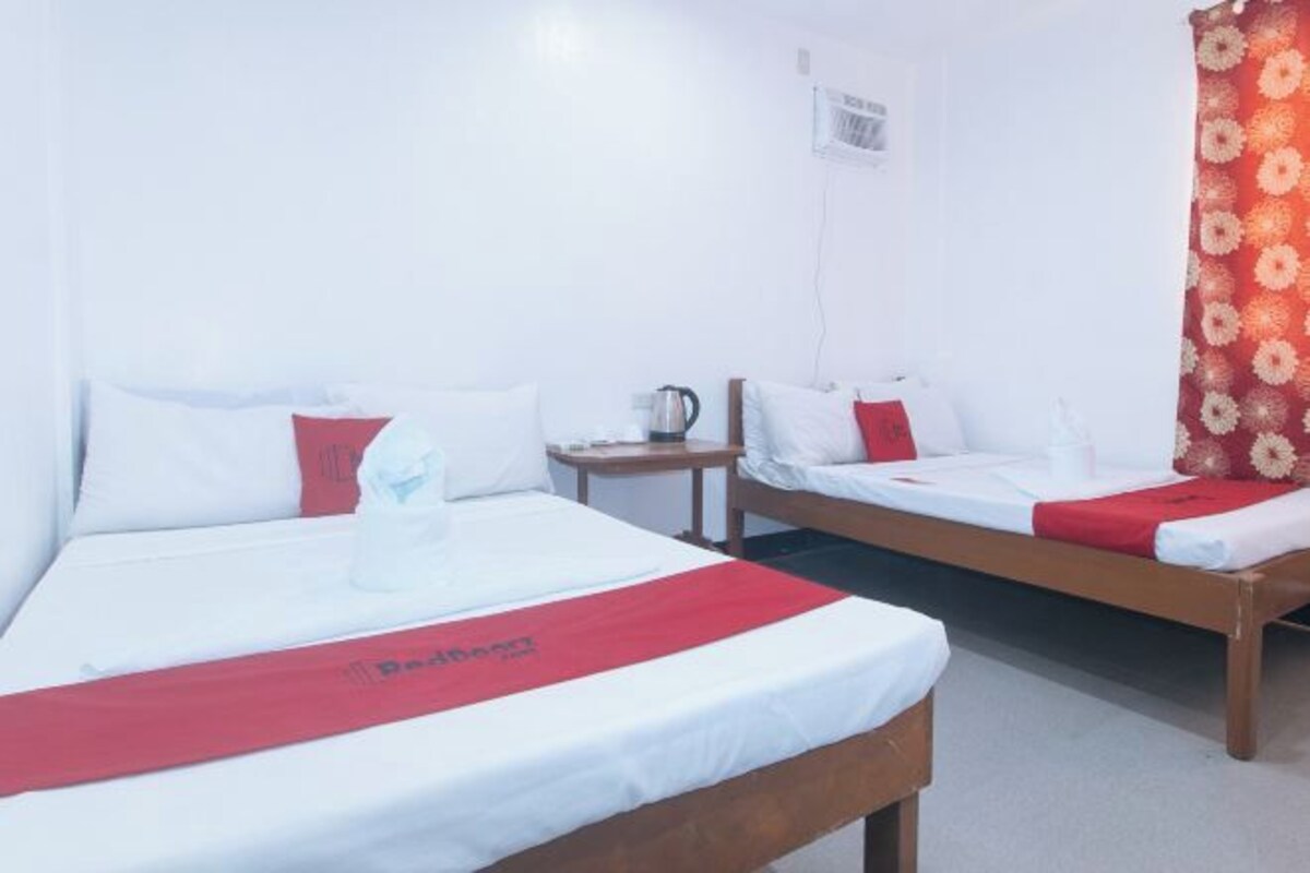 Een hotelkamer met twee bedden, elk voorzien van witte beddengoed en rode accenten, een klein tafeltje met een waterkoker en een raam met een bloemen gordijn.