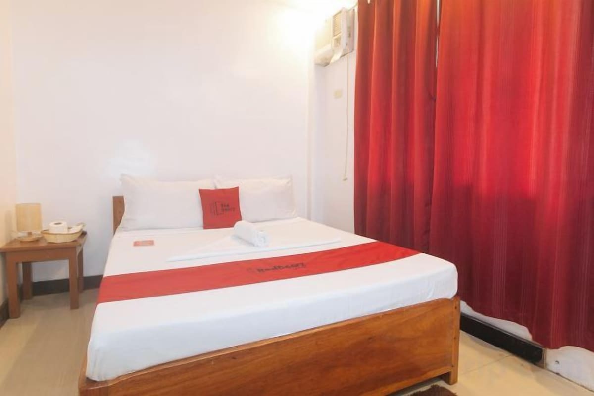 Ein Hotelzimmer mit einem hölzernen Bett, weißer Bettwäsche, einer roten Tagesdecke, einem Nachttisch mit Lampe und einem roten Vorhang.