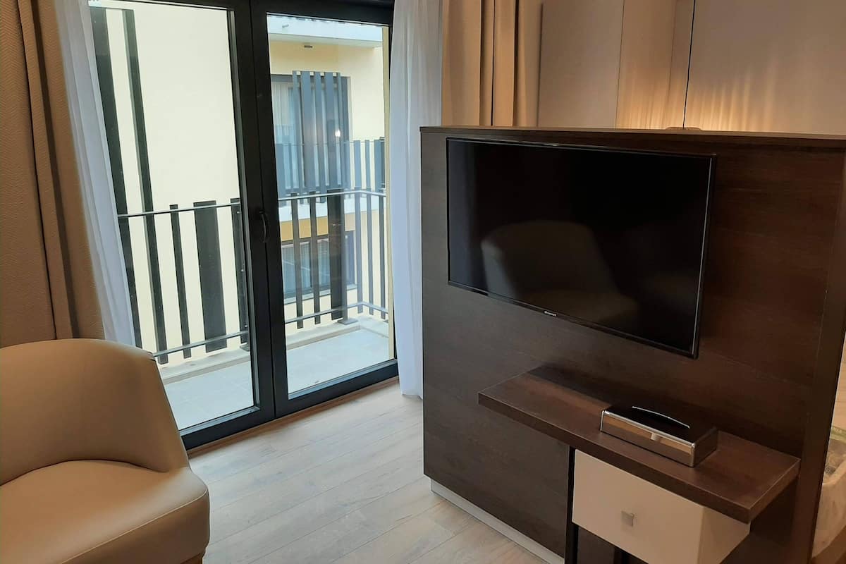 Een moderne hotelkamer met een grote tv gemonteerd op een donker houten paneel, een comfortabele beige fauteuil en een glazen schuifdeur naar een balkon met een balustrade.