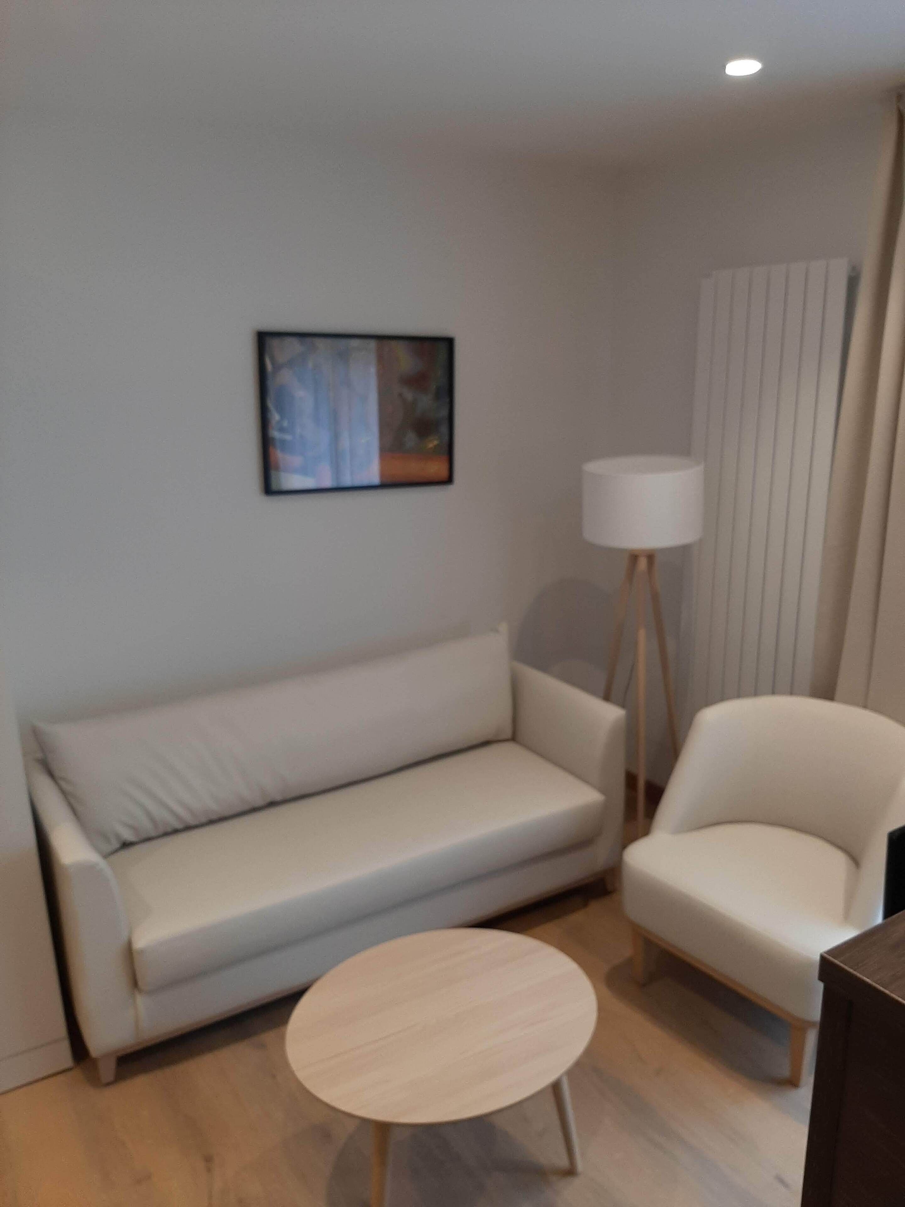 A modern living room with a sofa, a chair, a small round table, a floor lamp, and a painting on the wall.