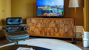 Een woonkamer met een televisie waarop een stadsgezicht te zien is, een zwarte leren fauteuil, een houten kast en een vloerlamp.