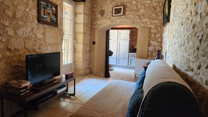 Une pièce aux murs de pierre, équipée d’une télévision, d’un canapé et offrant une vue sur la cuisine par une porte ouverte.