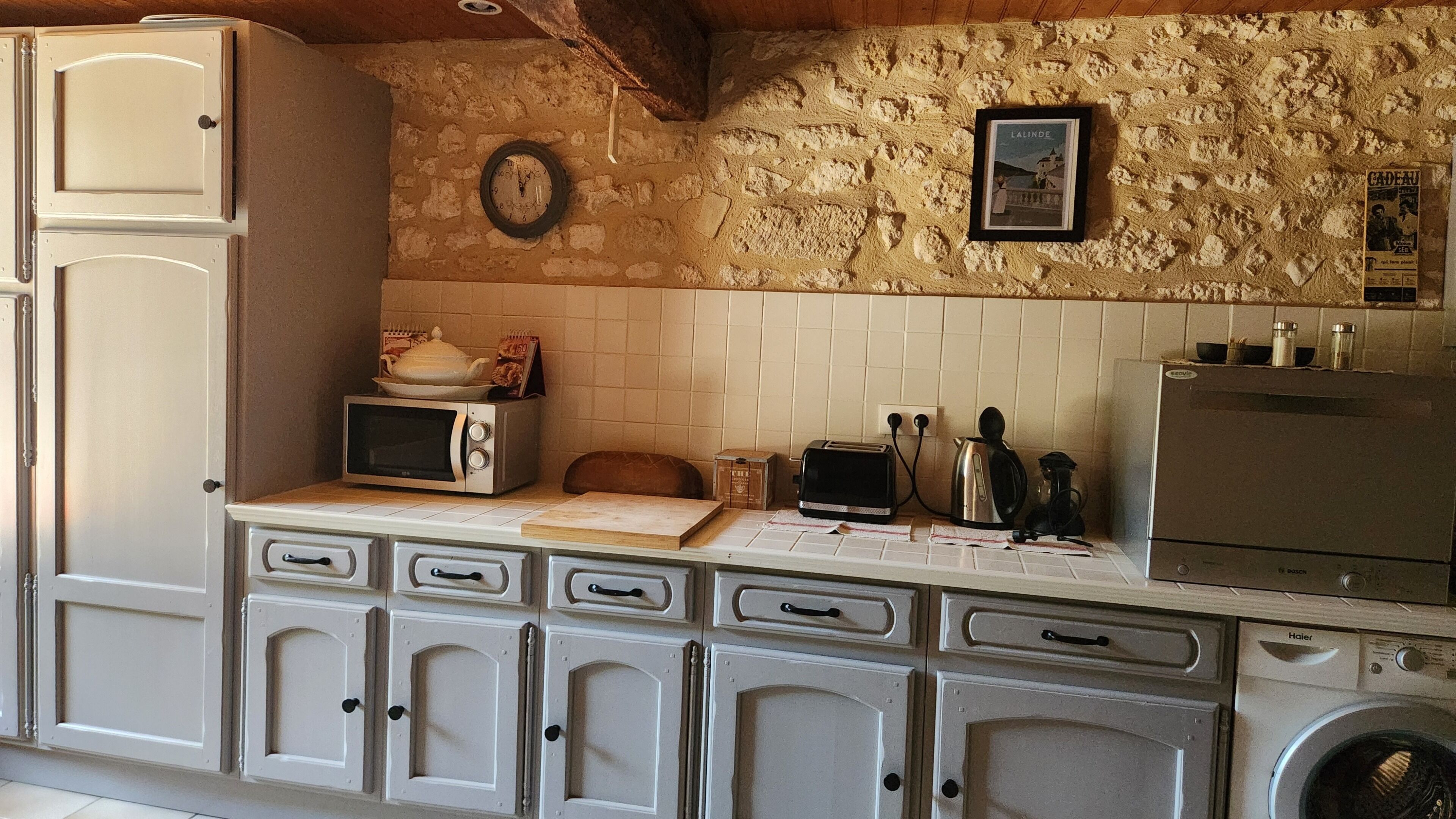 A kitchen with a stone wall, a clock, a framed picture, a microwave, a toaster, a kettle, and a washing machine.