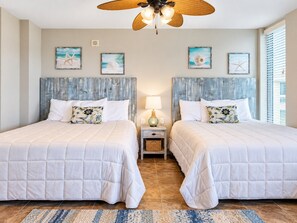 A bedroom with two beds, a ceiling fan, and artwork on the walls. - Beachfront Studio | Gulf Views, Balcony + Pools (Panama City Beach)