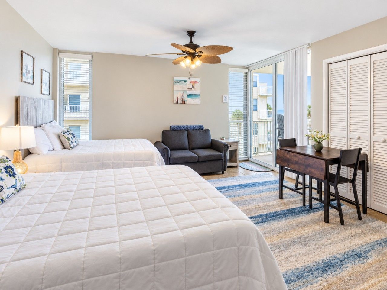 A hotel room with two beds, a ceiling fan, a desk with chairs, and a view of the outside.