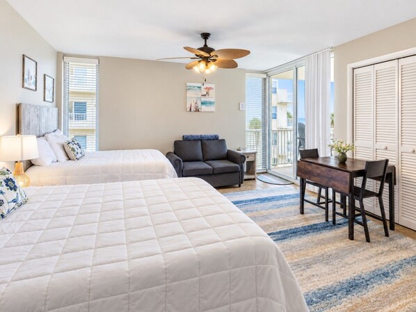 A hotel room with two beds, a ceiling fan, a desk with chairs, and a view of the outside.