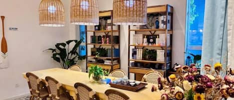 Un comedor con una mesa larga, sillas de mimbre y lámparas colgantes de mimbre. Hay estanterías con libros y objetos decorativos, y un jarrón con flores sobre la mesa.