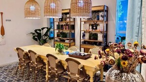 A dining area with a long table, wicker chairs, and hanging wicker lamps. There are bookshelves with books and decorative items, and a large window with a view. - Kela Coast Seaside Inn (Shenzhen)