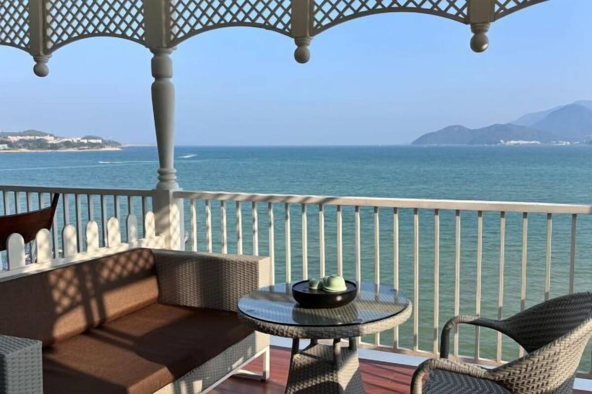 A balcony with wicker furniture, a round table, and a view of the sea and distant land.