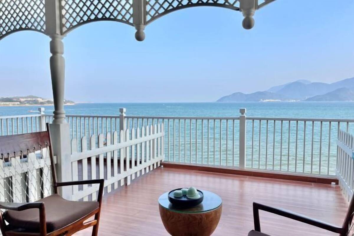 A balcony with a view of the sea, a wooden chair, a small table, and a white fence.