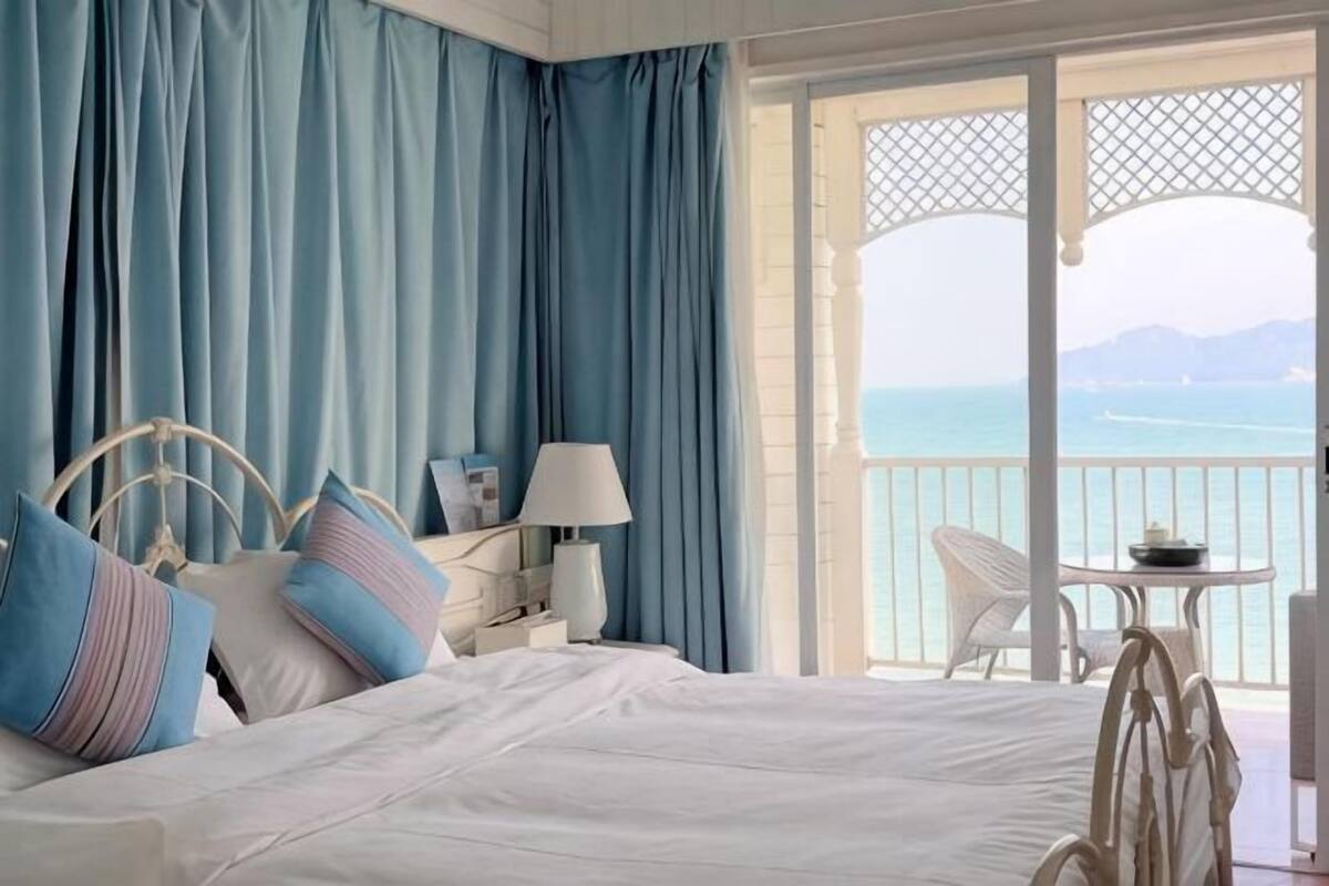 A bedroom with a bed, a nightstand, a lamp, a chair, and a view of the sea.
