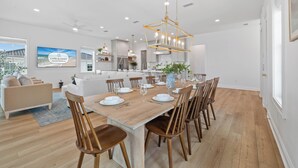 Une salle à manger spacieuse, avec une grande table en bois dressée pour six personnes, entourée de chaises en bois. Un lustre moderne est suspendu au plafond, et la pièce est agrémentée de murs blancs et d’un plancher en bois.