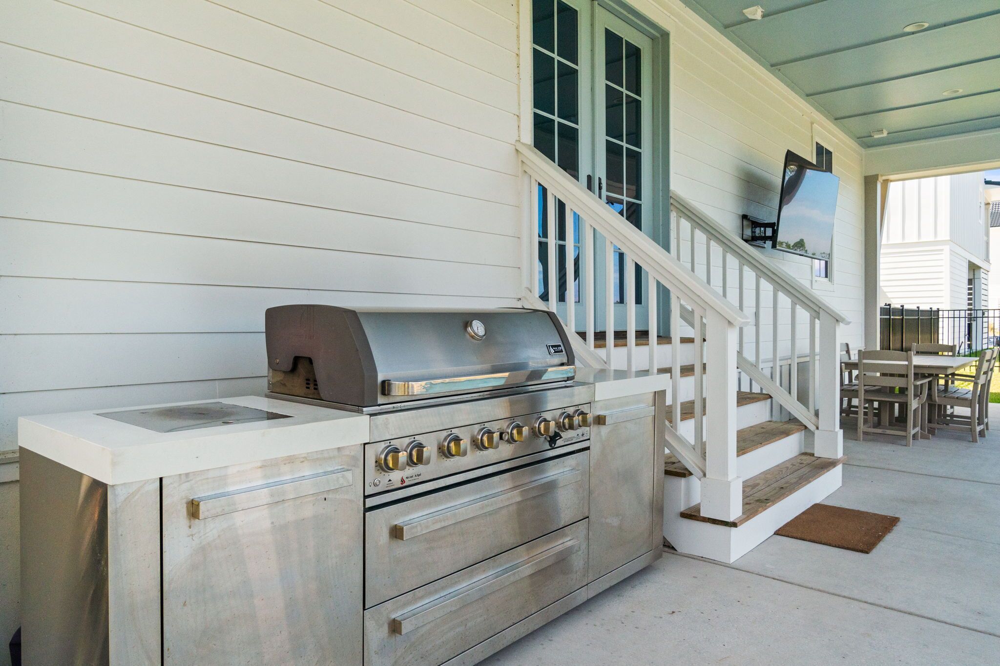 Una cocina exterior de acero inoxidable con parrilla, horno y pileta, al lado de un porche blanco con escaleras y comedor.
