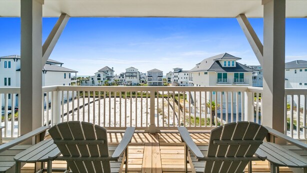 Un balcon agrémenté de deux fauteuils Adirondack et d’une table en bois, offrant une vue sur une plage et plusieurs maisons.
