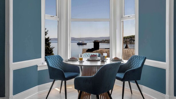 Un comedor con una mesa lista para dos, dos sillas azules y vista a un cuerpo de agua con un barco.