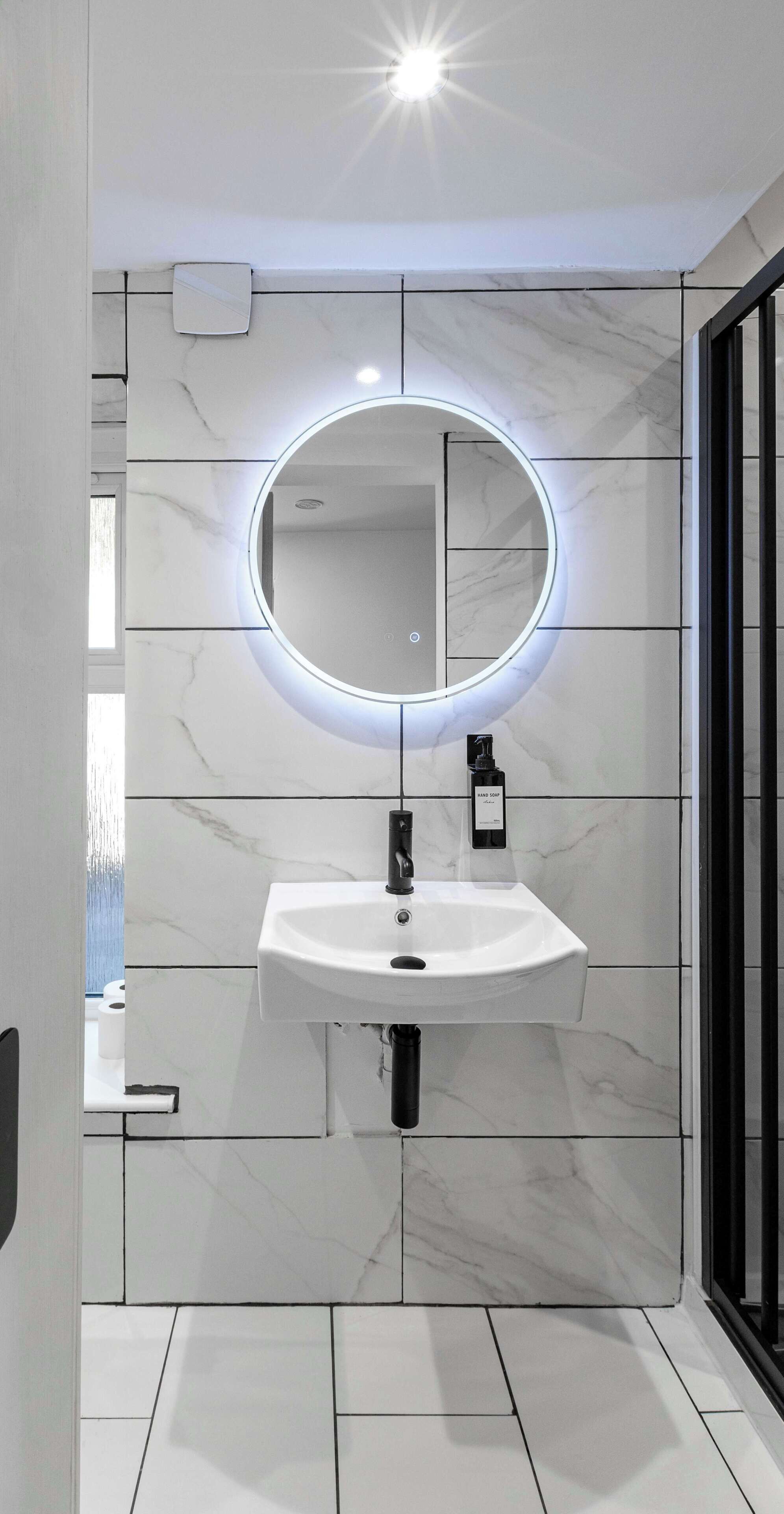A bathroom with a white marble sink, a round mirror with underglow lighting, and a black door.