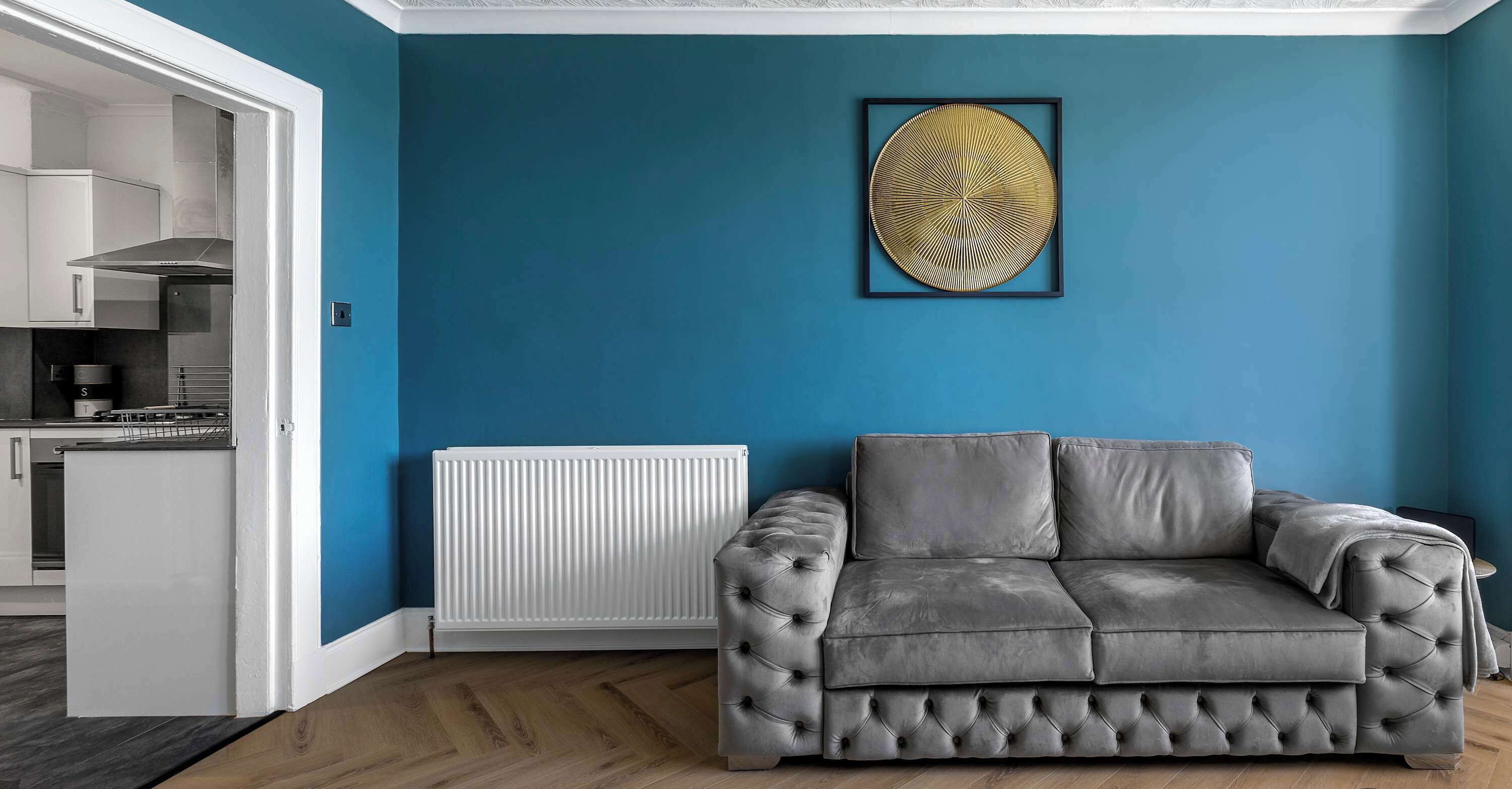 Una sala de estar con un sofá gris, un radiador blanco y una cocina visible a través de una puerta abierta.