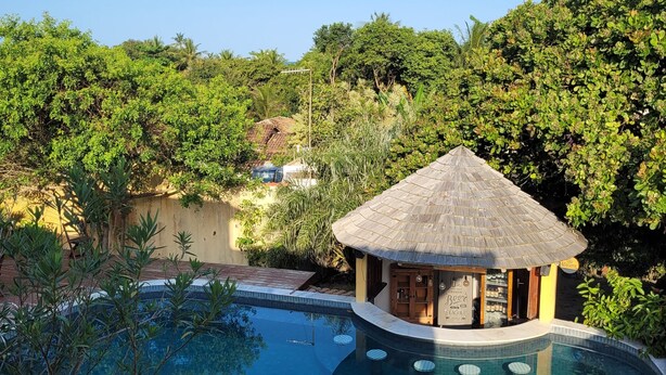A pool area with a thatched-roof structure, surrounded by lush greenery and a clear blue sky. - Pousada Oceano Caraiva (Porto Seguro)