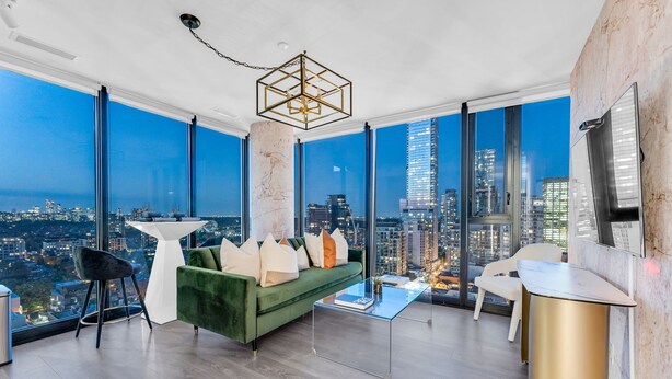A high-rise apartment with a city view, a green sofa, a glass coffee table, and a modern chandelier.