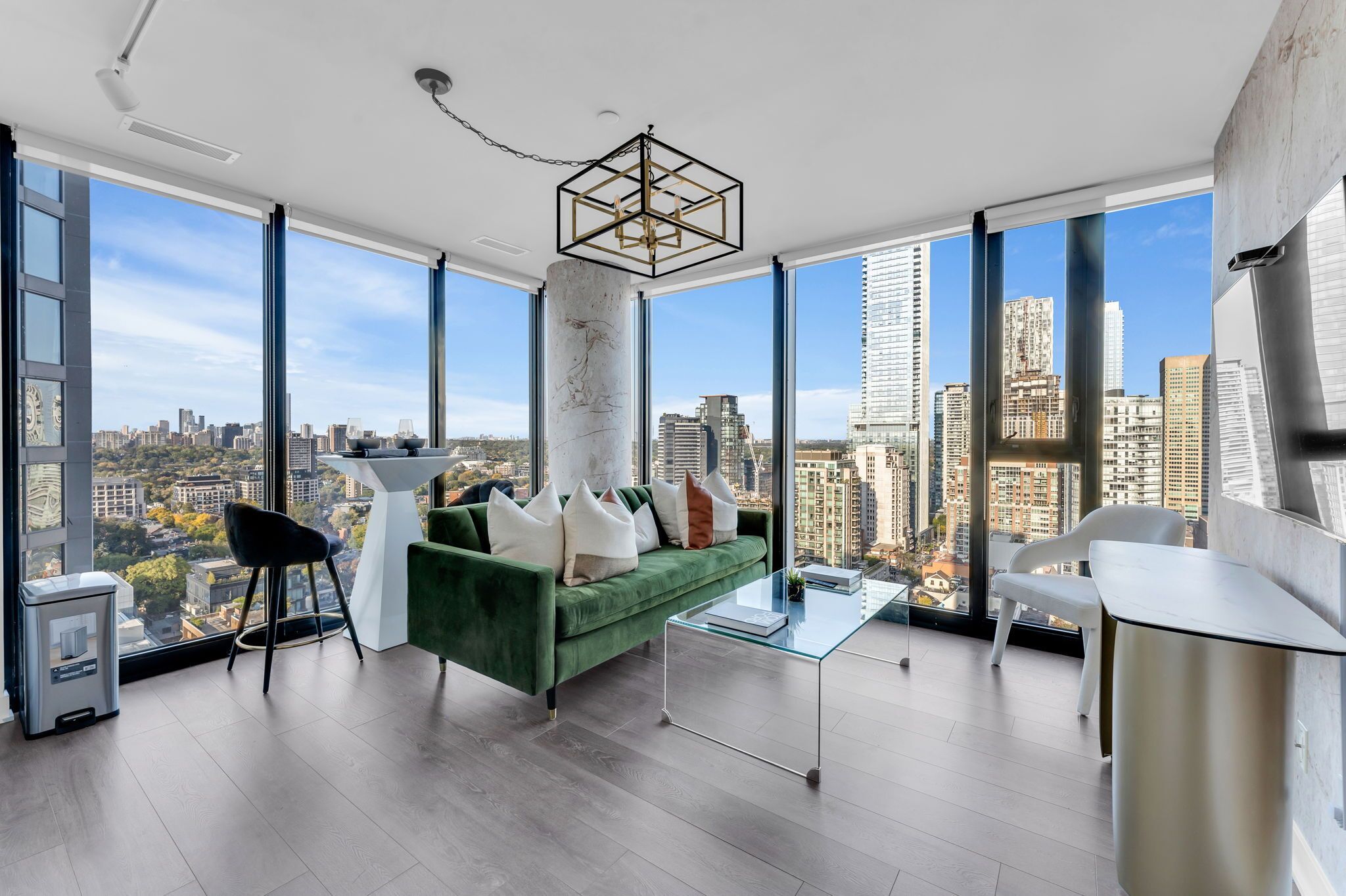 A spacious living room with a green sofa, a glass coffee table, and a view of a cityscape through large windows.