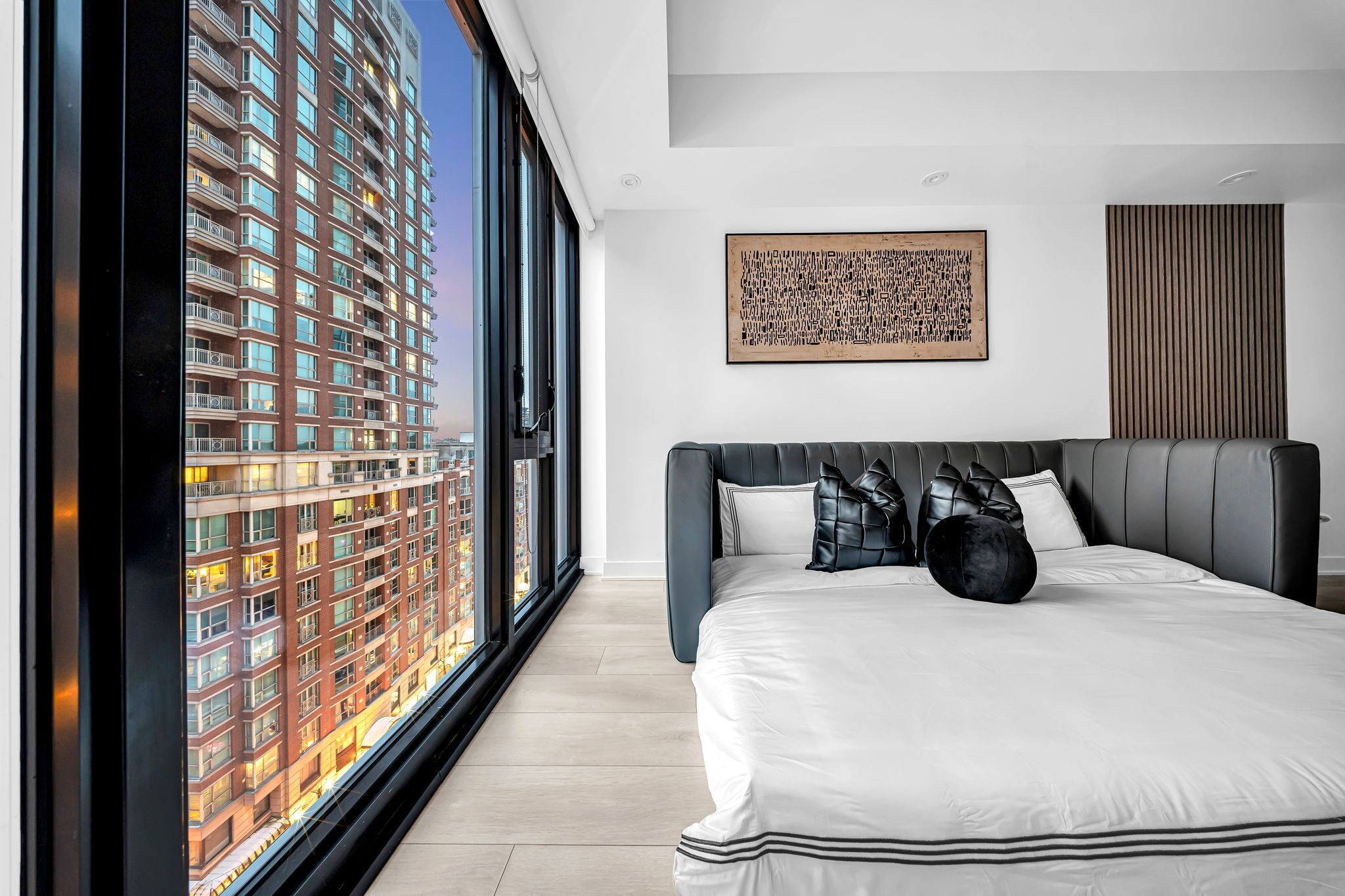 A modern bedroom with a large bed, a city view through floor-to-ceiling windows, a wooden panel wall, and a framed artwork.