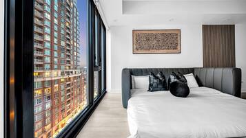 A modern bedroom with a large bed, a city view through floor-to-ceiling windows, a wooden panel wall, and a framed artwork.