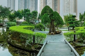 A wooden pathway leading to a pond with a large tree in the center, surrounded by neatly trimmed bushes and tall residential buildings in the background. - Dongguan Junlan Hotel (Houjie Wanda Plaza) (Dongguan)