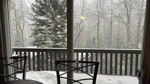Une vue depuis une fenêtre donnant sur une terrasse en bois et des chaises, avec en contrebas un paysage enneigé parsemé d’arbres.