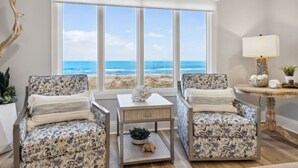 Ein Zimmer mit großem Fenster mit Blick auf einen Strand, zwei Sessel mit Blumenmuster, ein kleiner Tisch mit Lampe und eine Zierpflanze.