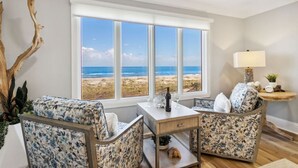 Ein Zimmer mit großem Fenster mit Blick auf einen Strand, zwei stuhlähnliche Sitzgelegenheiten mit Blumenmuster, ein kleiner Tisch mit Weinflasche und Gläsern sowie eine Lampe auf einem Beistelltisch.