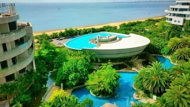Ein Hotel mit einem kreisförmigen Pool, umgeben von üppiger Vegetation, Blick auf einen Strand und einem klaren Himmel.