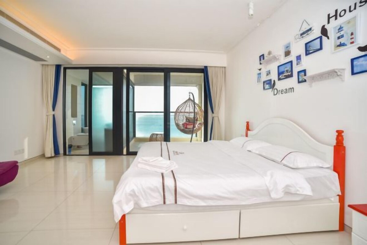 A hotel room with a bed, a view of the sea through a glass door, and a wall with framed pictures and the word "Dream".