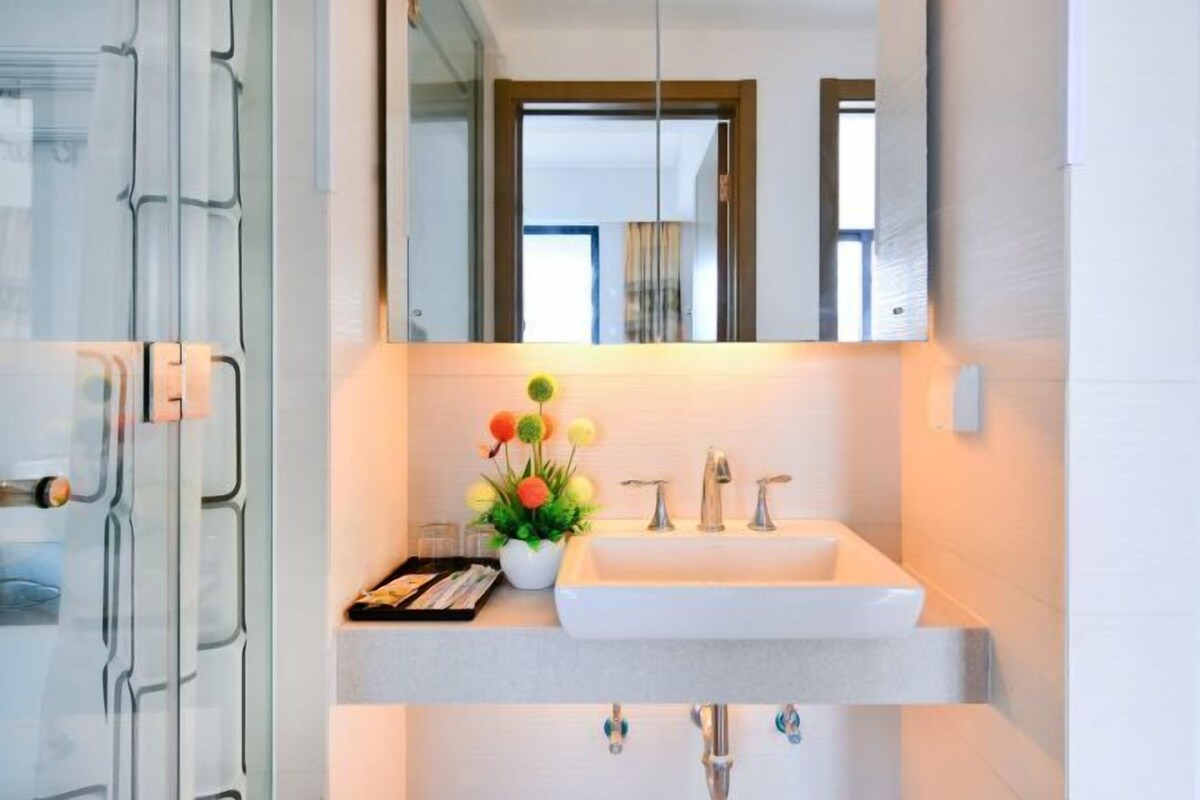 A modern bathroom with a large mirror, a white sink, and a small flower arrangement on the countertop.