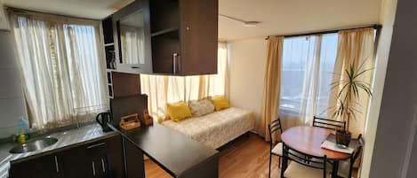 A compact hotel room with a kitchenette, a bed with yellow pillows, a dining table with chairs, and a potted plant by the window.