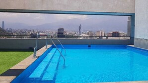 Ein Pool auf dem Dach mit Blick auf die Stadtlandschaft, eine Metallleiter und eine kleine Poolanlage mit Rasenfläche.