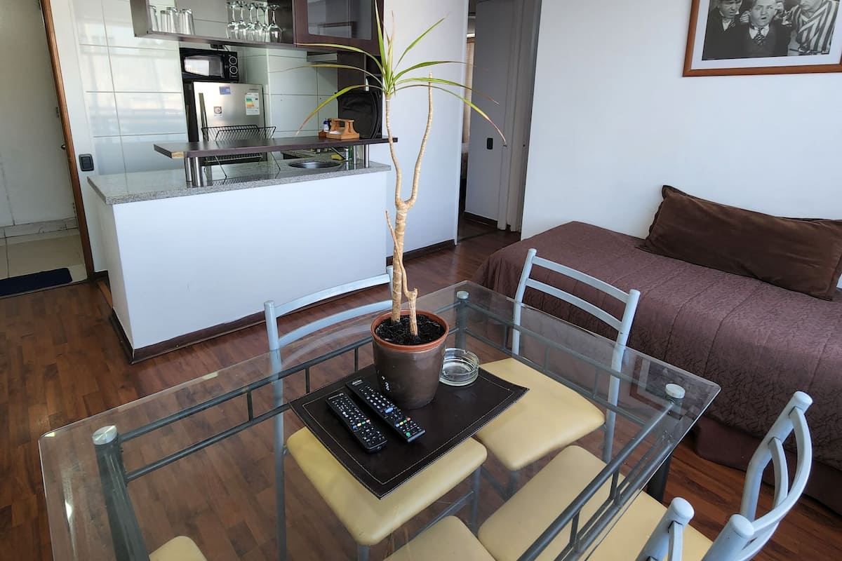 Un espacio habitable moderno con una mesa de vidrio, sillas, una planta en maceta, un sofá y una cocina con microondas y armarios.