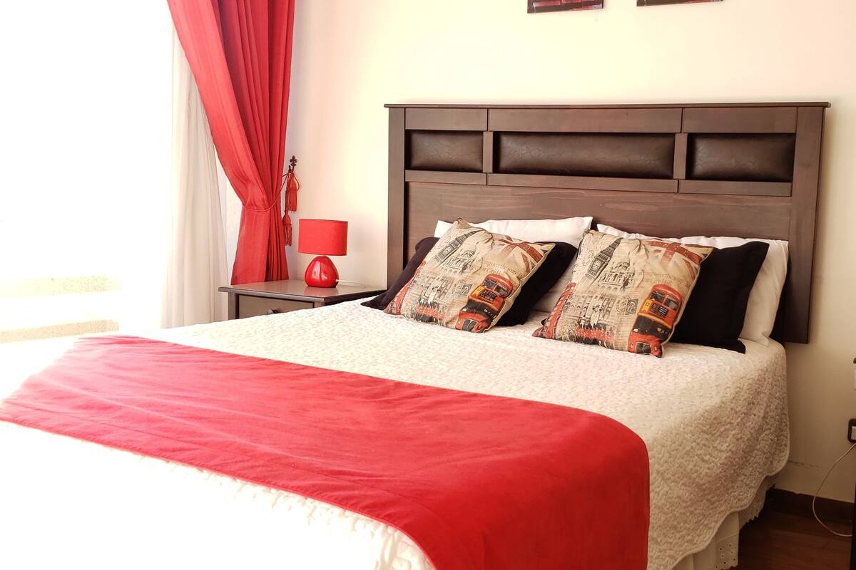 Una cama bien hecha con una colcha roja, dos lámparas de noche rojas y una mesita de noche con un control remoto.