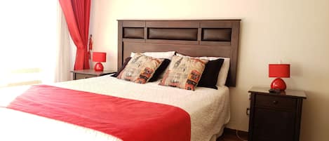 A neatly made bed with a red blanket, two red bedside lamps, and a small nightstand with a remote control.