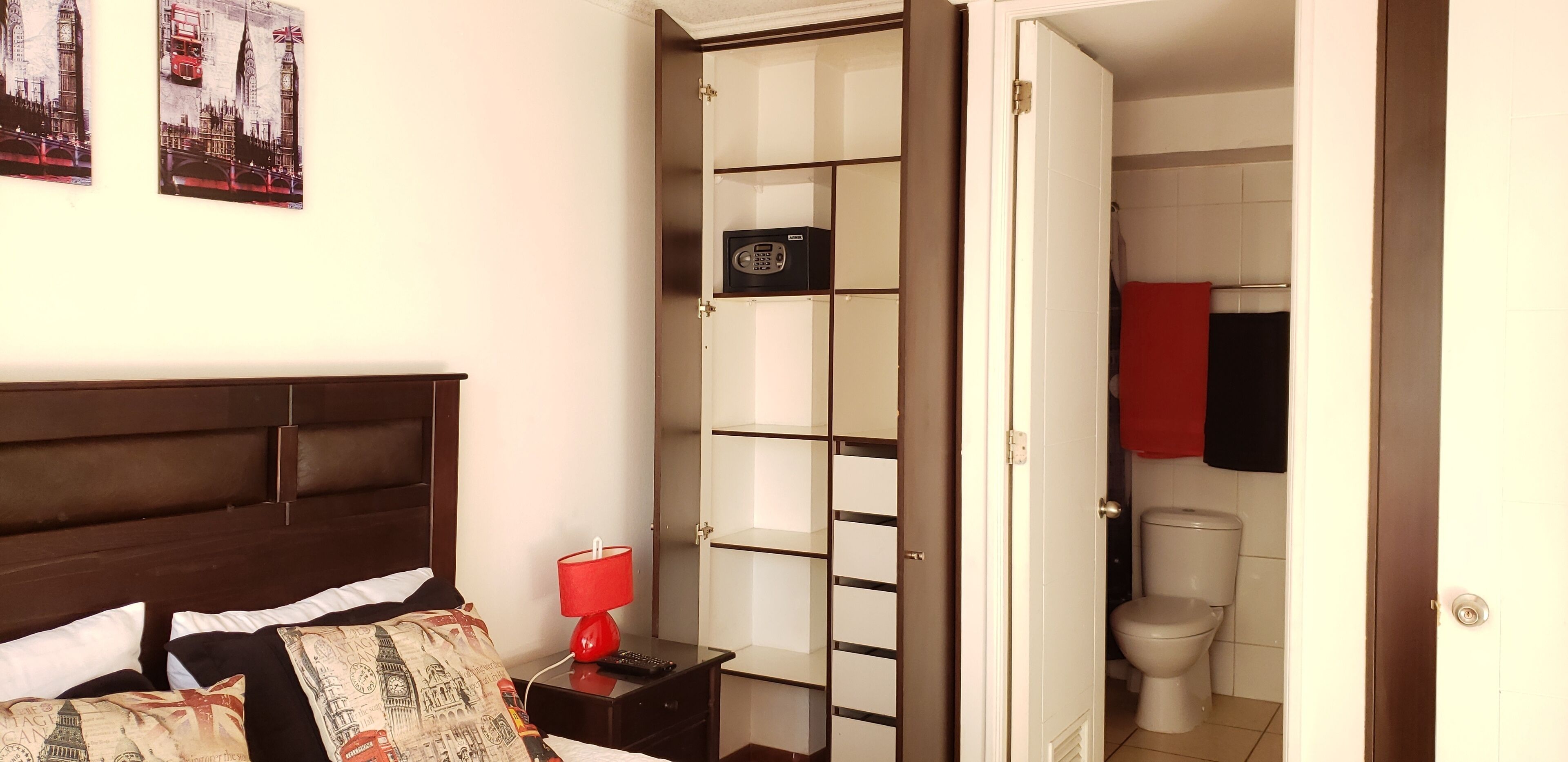 Habitación de hotel con cama, mesita de noche con lámpara, armario y baño visible a través de una puerta abierta.