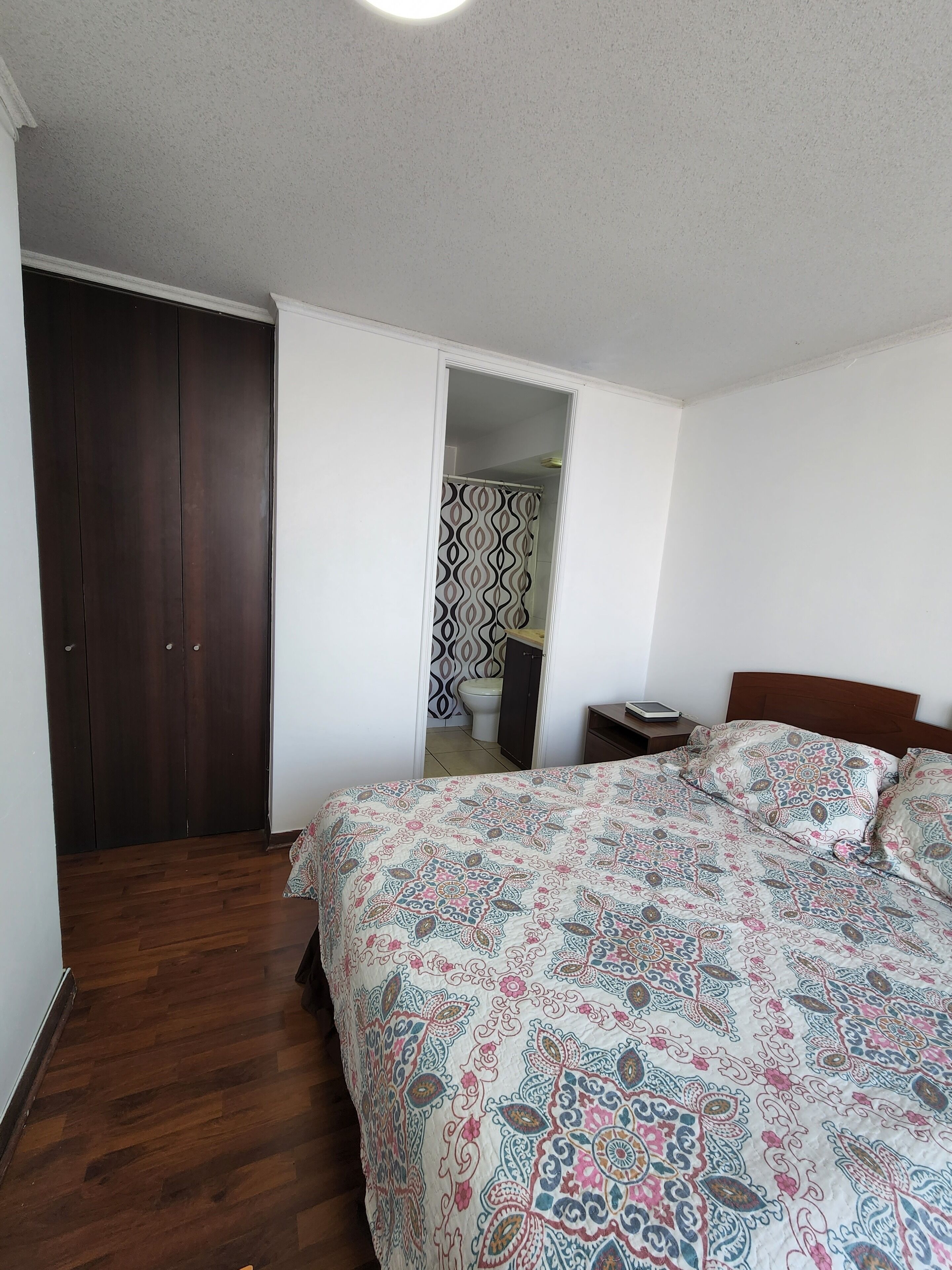 A bedroom with a wooden wardrobe, a bed with patterned bedding, a bedside table, and a view into the bathroom.