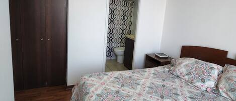 Un dormitorio con un armario de madera, una cama con ropa de cama estampada, una mesita de noche y vistas al baño.
