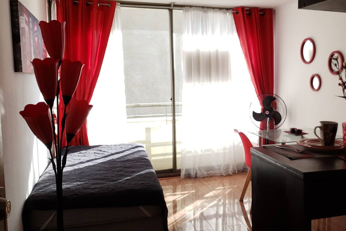 Una habitación con cama, una mesa de comedor con vasos rojos y un ventilador, y una ventana con cortinas rojas.